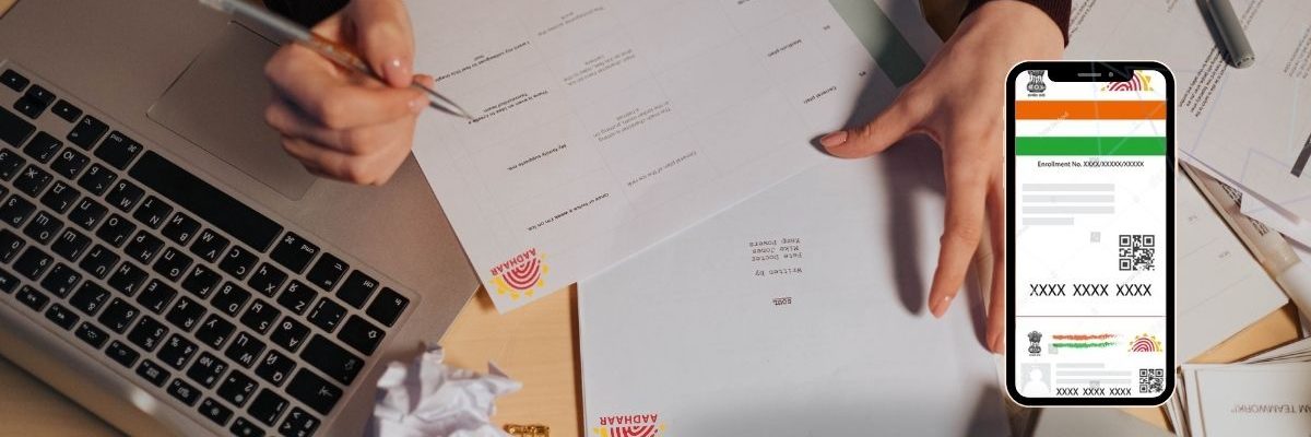Document Processing An overhead view of a person with a pen, signing and reviewing documents on a wooden desk. A crumpled piece of paper lies next to a laptop. On the right side, a smartphone displays the digital version of an Aadhar card, complete with an enrollment number, QR code, and other details. The documents on the table have an "AADHAAR" logo printed on them.